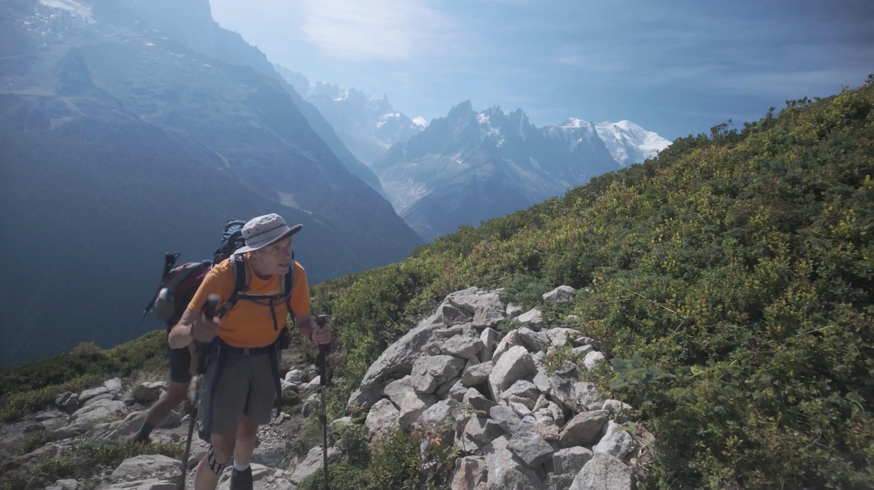 A hiker climbing a rocky mountain trail with alpine peaks in the distance